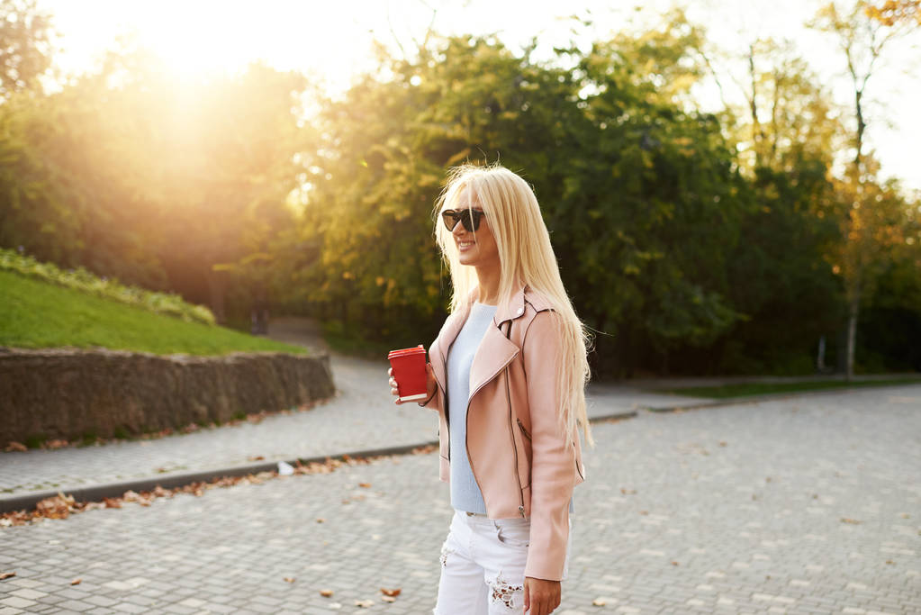 Het concept van street fashion. stijlvolle meisje student wandelen in het zonnige park en in het bezit van een kopje koffie te gaan. Portret van glimlachen blond meisje in zonnebril - Foto, afbeelding
