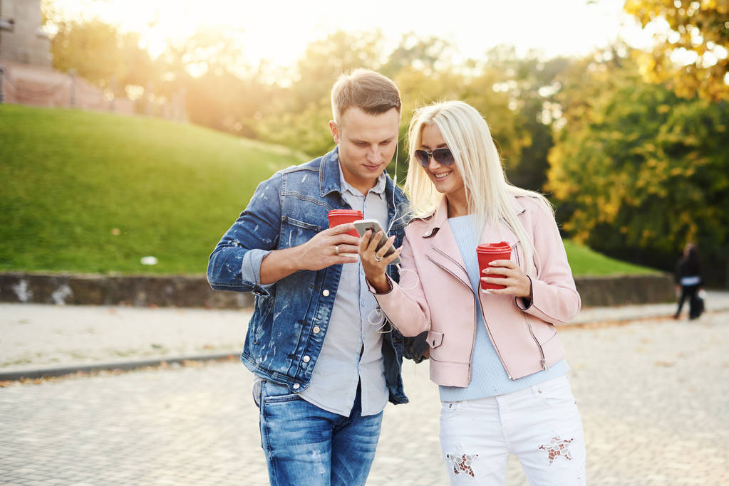 Hymyilevä pariskunta kävelee syksyllä puistossa kädestä pitäen. Kuuntele musiikkia kuulokkeilla ja juo kahvia mukaan. Pidä hauskaa yhdessä. Halaus ja suutelu
  - Valokuva, kuva
