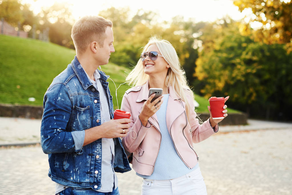 Улыбающаяся влюбленная пара, гуляющая в осеннем парке, держащаяся за руки. Слушать музыку в наушниках и пить кофе с собой. Веселитесь вместе. Объятия и поцелуи
  - Фото, изображение