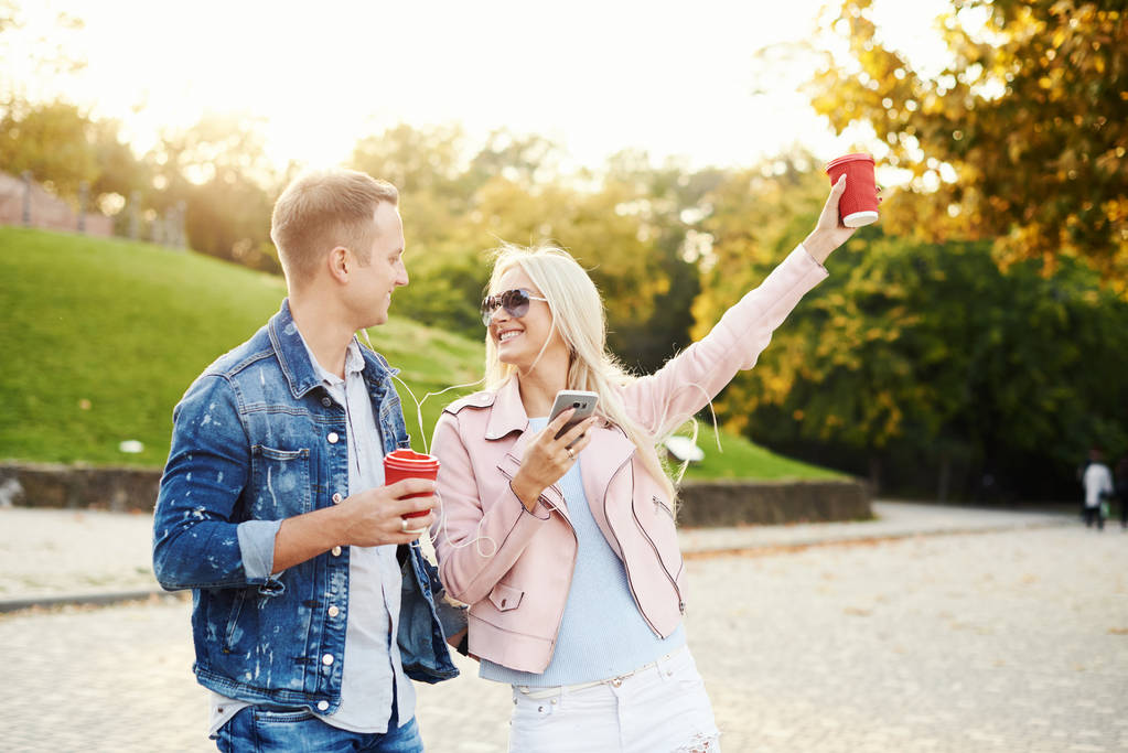 Usmívající se pár v lásce chůze v podzimním parku, drželi se za ruce. Poslouchat hudbu z sluchátka a pít kávu s sebou. Bavte se společně. Objímání a líbání - Fotografie, Obrázek