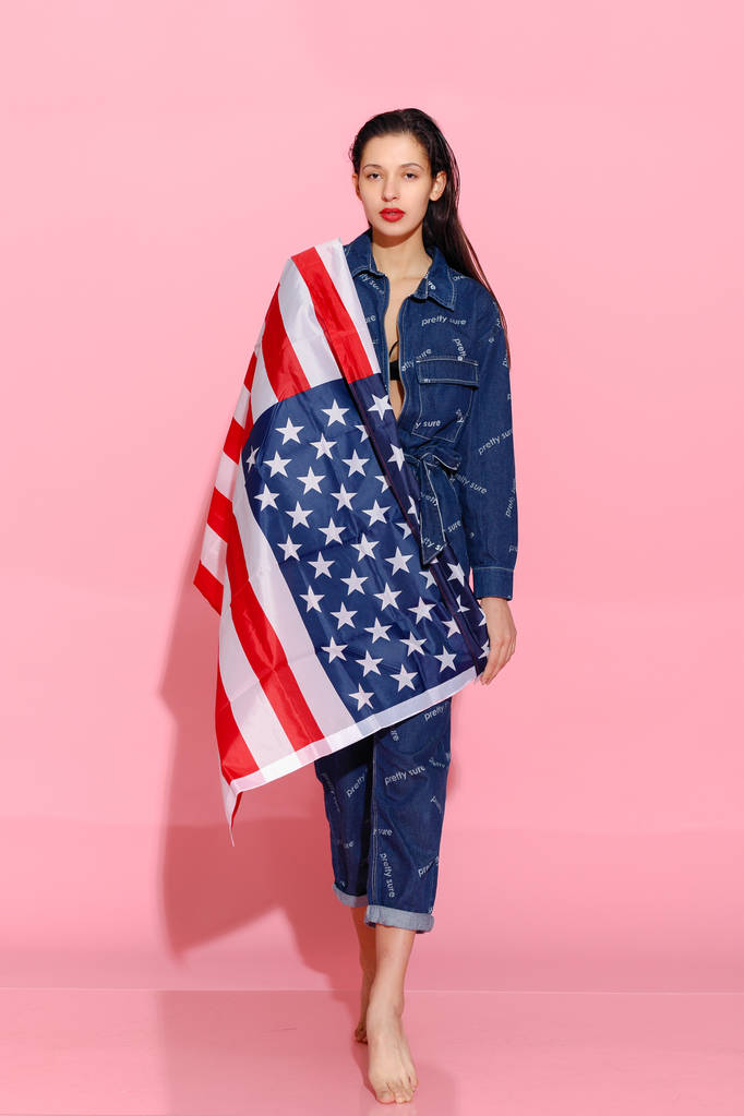 Portrait of proud female athlete wrapped in American Flag against pink background. Muscular young woman looking confidently at camera. female protesting for equality and women empowerment - Photo, Image