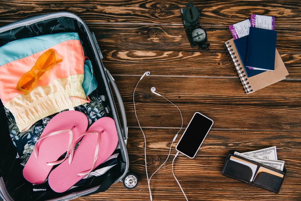 felülnézete a fa felülettel smartphone-val fülhallgató, iránytű, útlevelek jegyek és a nyári ruhák az utazótáska - Fotó, kép