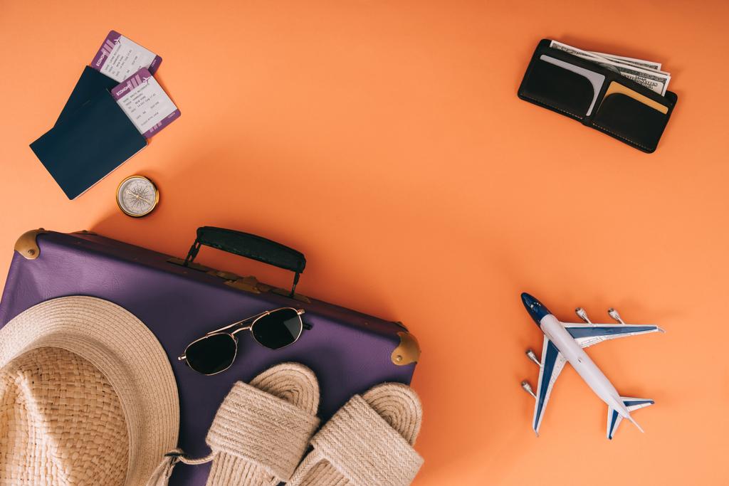 вид сверху летних аксессуаров на дорожную сумку, модель самолета, бумажник и паспорта с билетами на оранжевом фоне
 - Фото, изображение