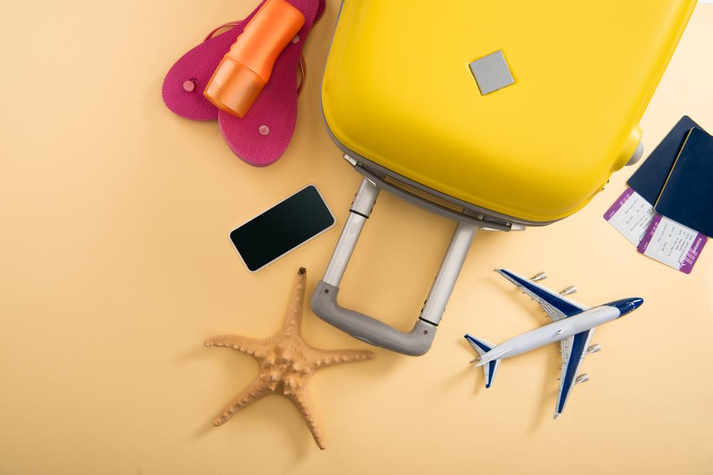Draufsicht auf gelben Koffer, Flugzeugmodell, Seestern, Sonnencreme, Flip-Flops, Smartphone und Tickets auf beigem Hintergrund - Foto, Bild