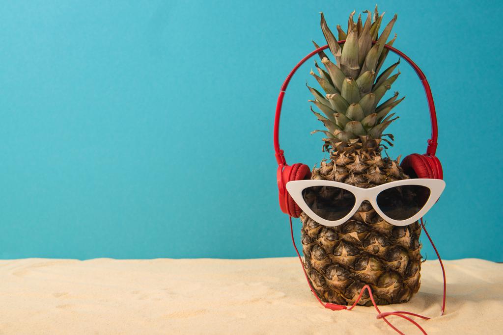 Ananas mit Kopfhörern und Sonnenbrille auf Sand auf blauem Hintergrund - Foto, Bild