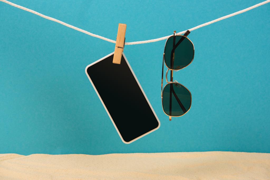 Smartphone mit leerem Bildschirm und Sonnenbrille am Seil mit Pin auf blauem Hintergrund - Foto, Bild