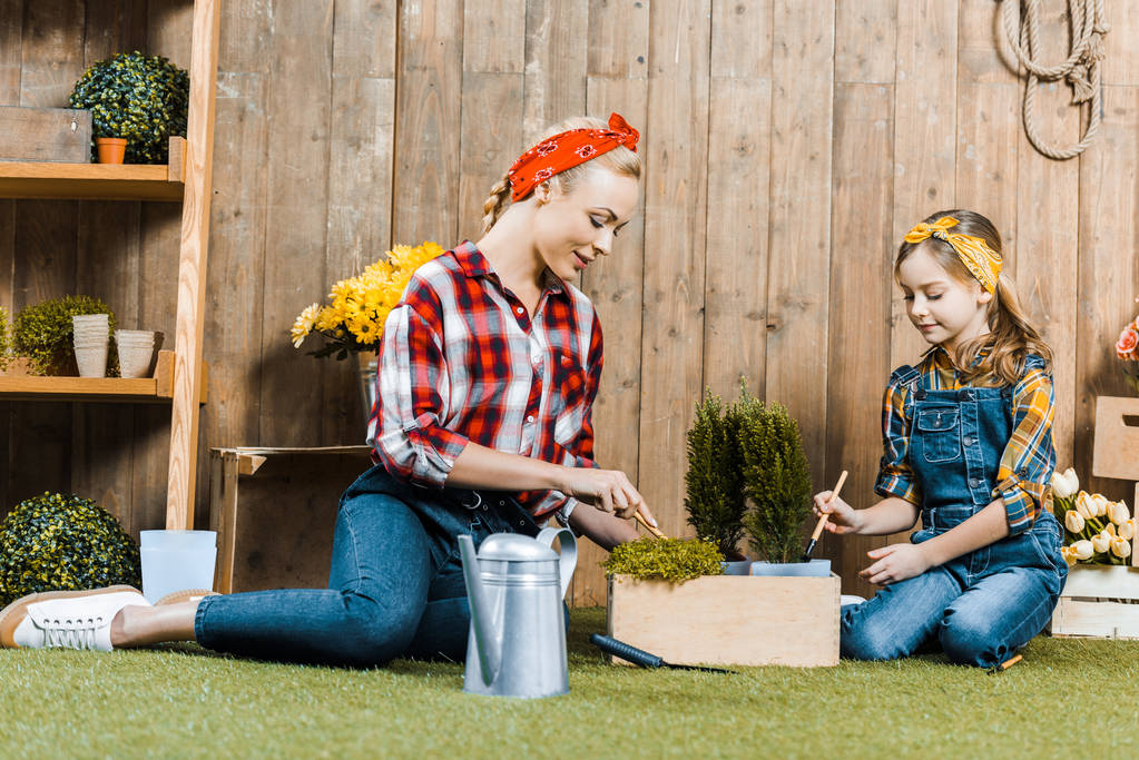 mother planting flowers near daughter while sitting on grass near wooden fence - Photo, Image