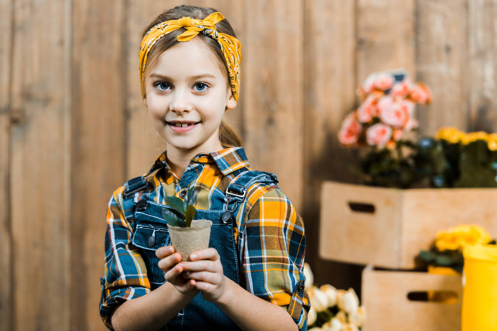 niño feliz sosteniendo una pequeña planta con hojas verdes cerca de la cerca
 - Foto, Imagen
