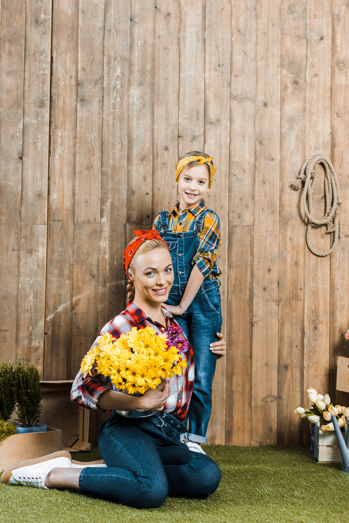 fröhliche Frau hält Blumen in der Hand und sitzt neben Tochter auf Gras - Foto, Bild
