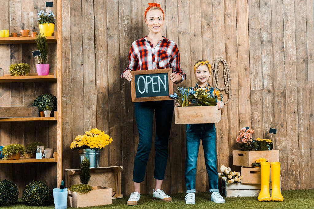 atraktivní žena držící malé tabuli s otevřenou nápisy poblíž roztomilý dcera drží box s rostlinami - Fotografie, Obrázek