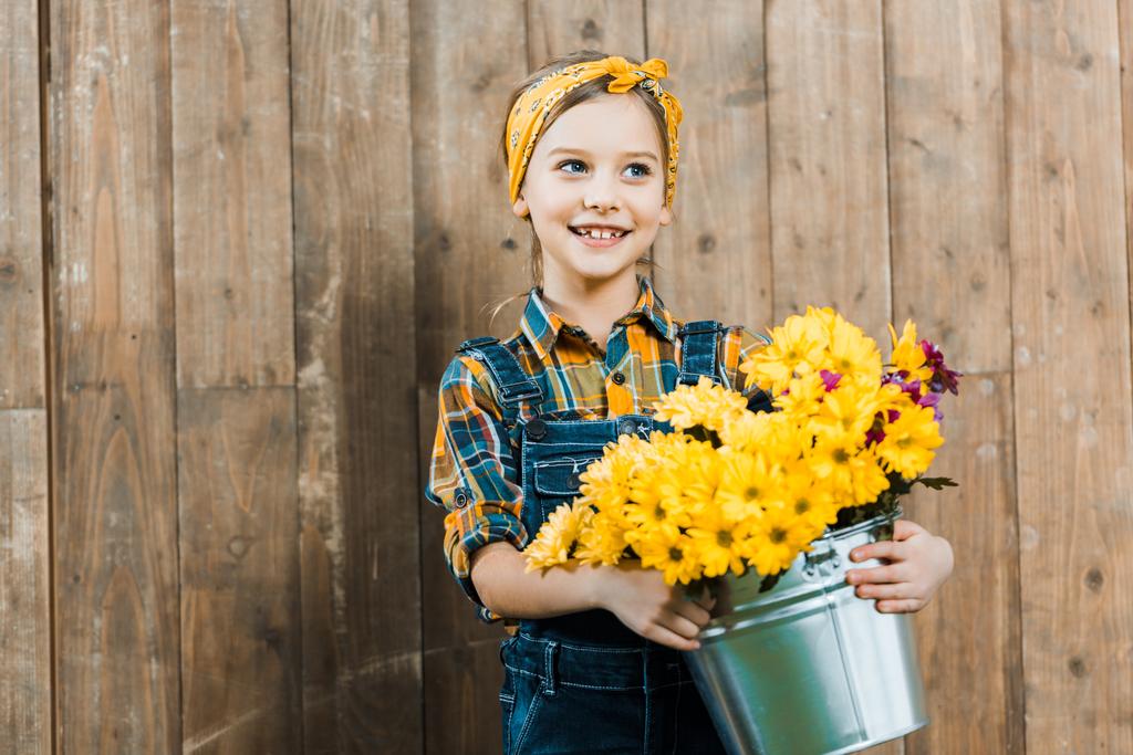 niño alegre sosteniendo flores en cubo y de pie cerca de valla de madera
 - Foto, imagen