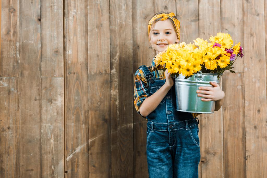 criança feliz segurando flores em balde e de pé perto de cerca de madeira
 - Foto, Imagem
