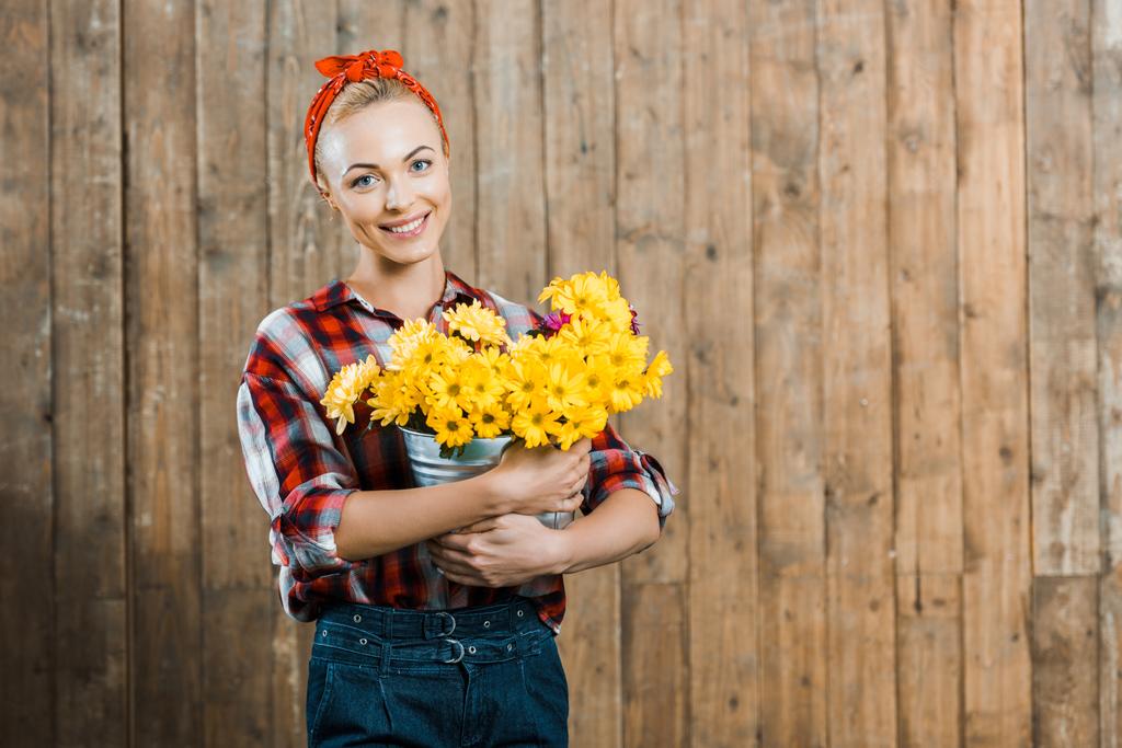 bela mulher segurando flores em balde e sorrindo perto de cerca de madeira
 - Foto, Imagem