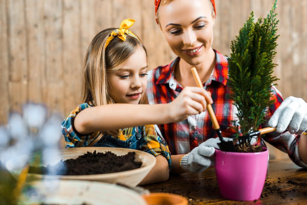 beautiful mother and cute daughter holding small rakes near ground with plant in pot - Photo, Image