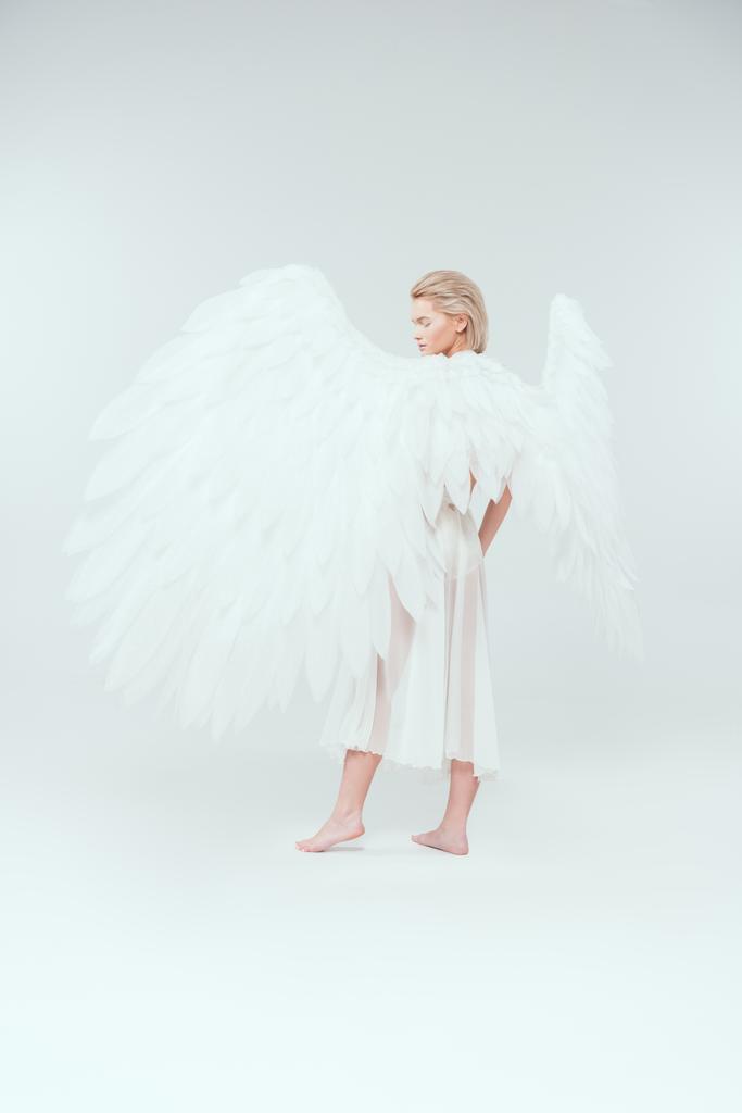όμορφη νεαρή γυναίκα με άγγελο φτερά αποστρέφεις το βλέμμα και ποζάρει απομονωθεί σε λευκό - Φωτογραφία, εικόνα