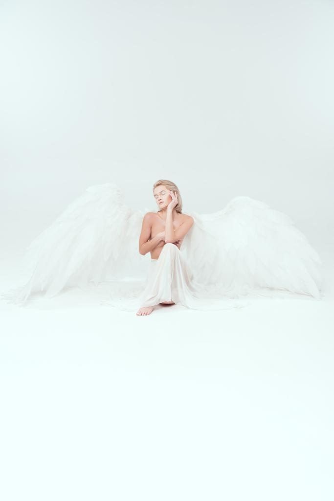 όμορφη γυναίκα με φτερά αγγέλου που καλύπτει το στήθος και ποζάρουν απομονωθεί σε λευκό με αντίγραφο χώρου - Φωτογραφία, εικόνα