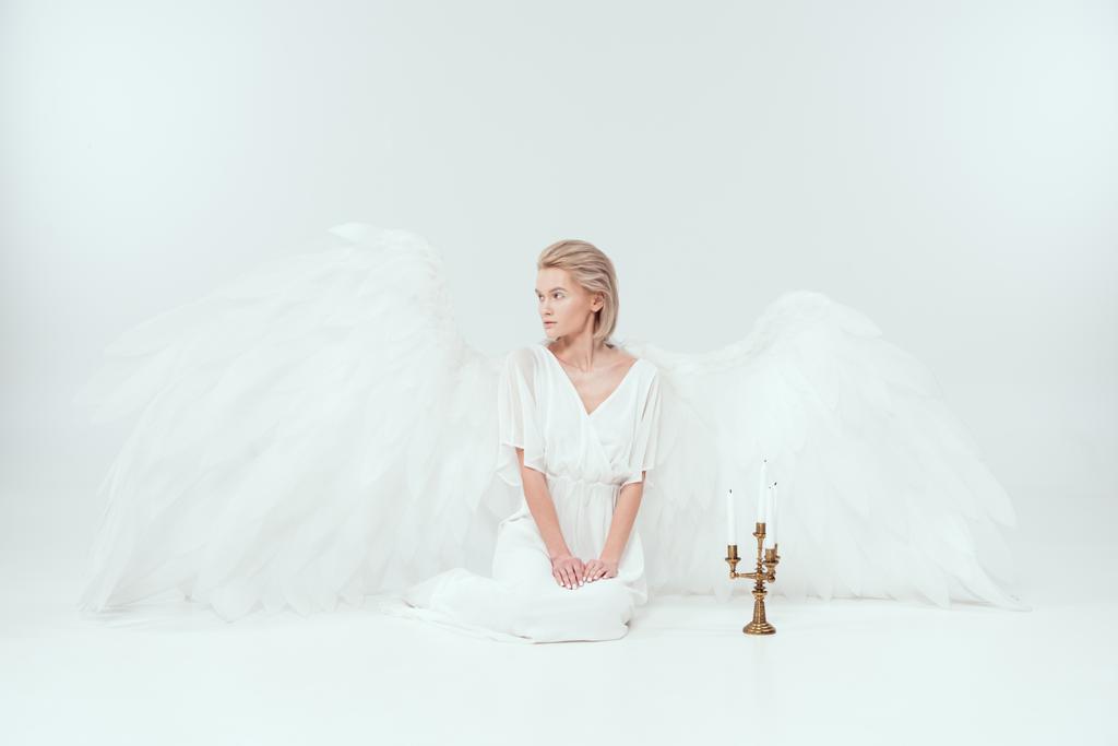 hermosa mujer en traje de ángel con alas sentadas cerca de candelabro con velas y mirando hacia otro lado aislado en blanco
 - Foto, Imagen