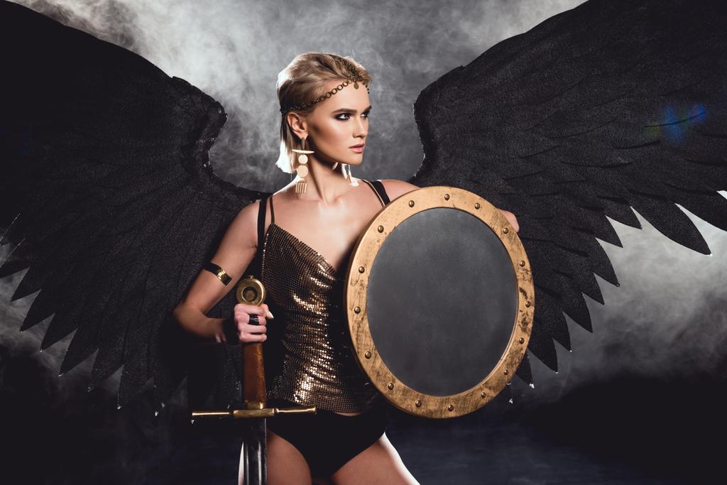 Krásná sexy žena bojovník kostým a angel wings pózuje s štít a meč na černém pozadí - Fotografie, Obrázek