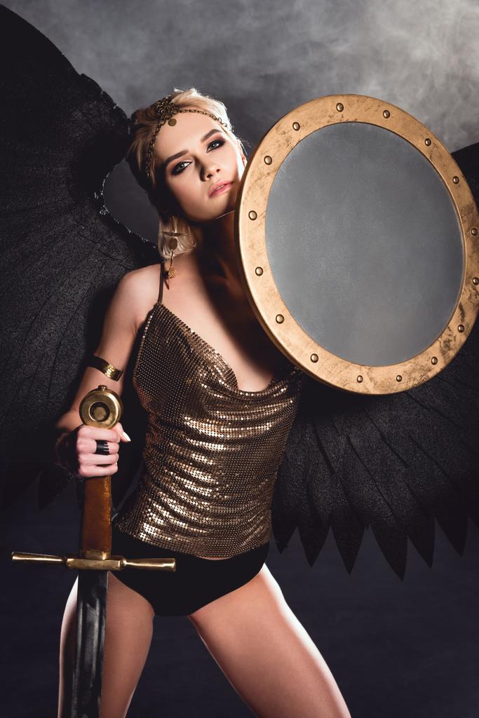 beautiful sexy woman in warrior costume and angel wings posing with shield and sword on dark background - Photo, Image