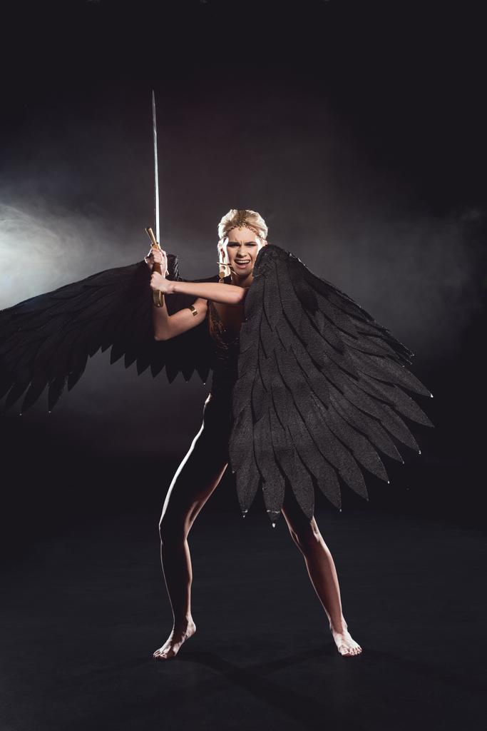 όμορφη θυμωμένη γυναίκα στο κοστούμι πολεμιστής με φτερά αγγέλου κρατάει σπαθί και θέτοντας σε μαύρο φόντο - Φωτογραφία, εικόνα