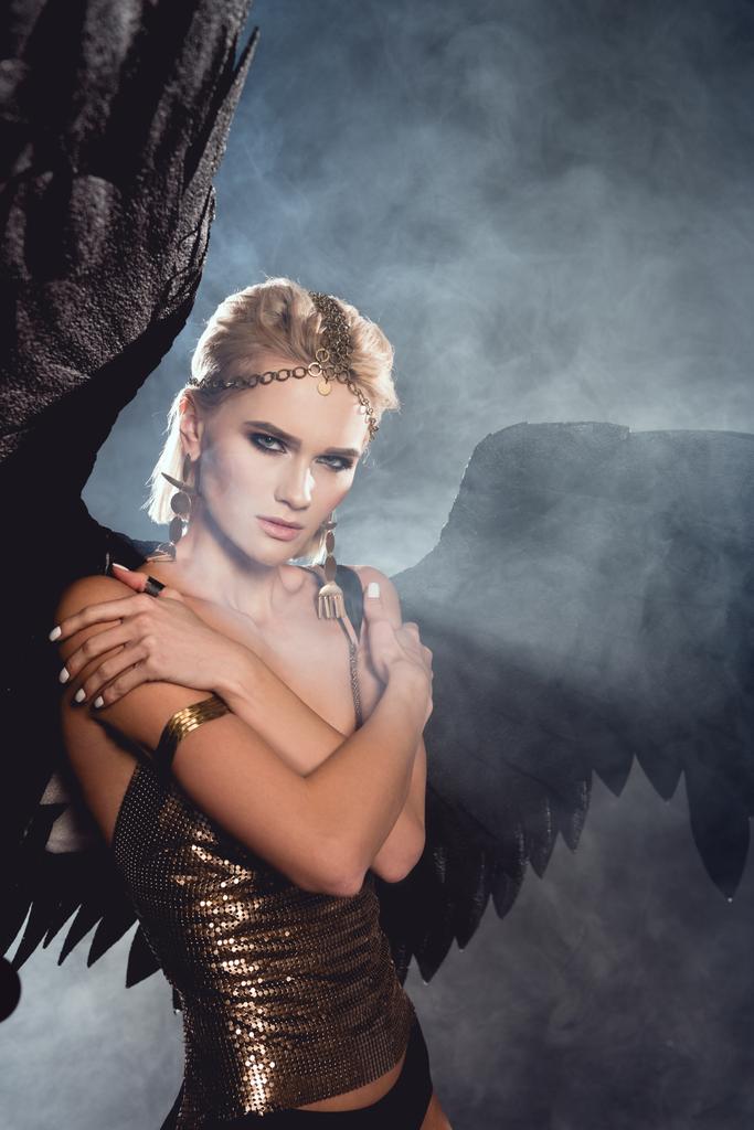beautiful young sexy woman with black angel wings and golden accessories posing on dark smoky background - Photo, Image