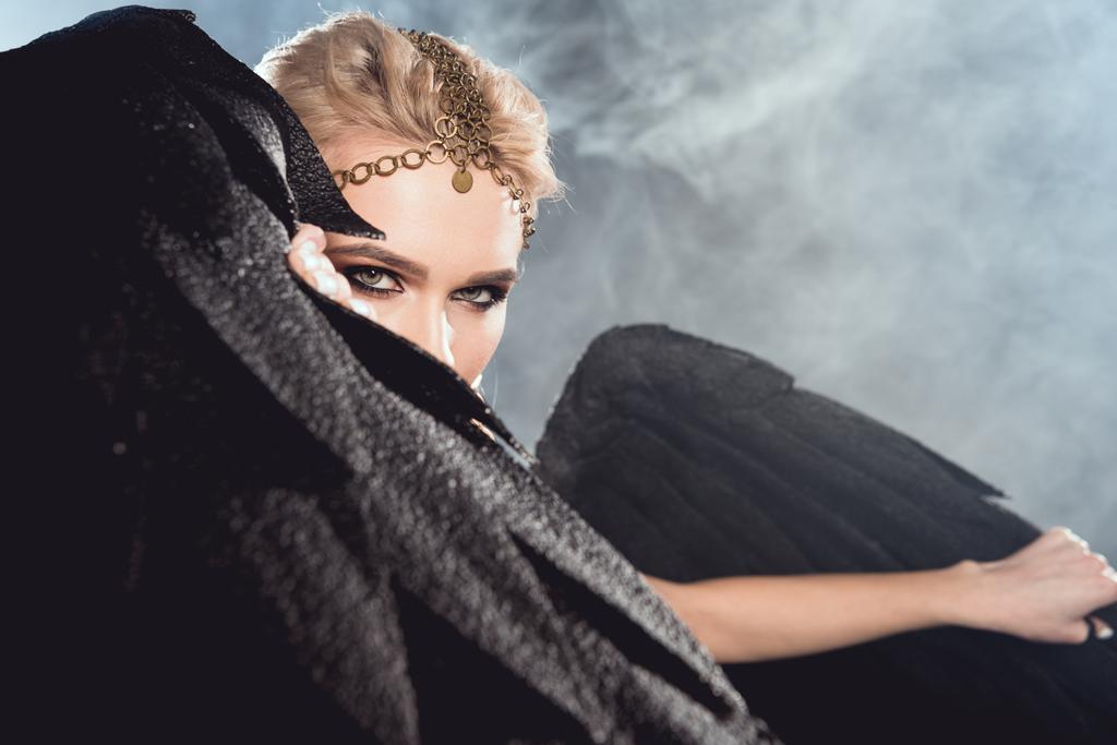 красивая сексуальная женщина в золотых аксессуарах держа крылья черного ангела и позируя на темном фоне
 - Фото, изображение