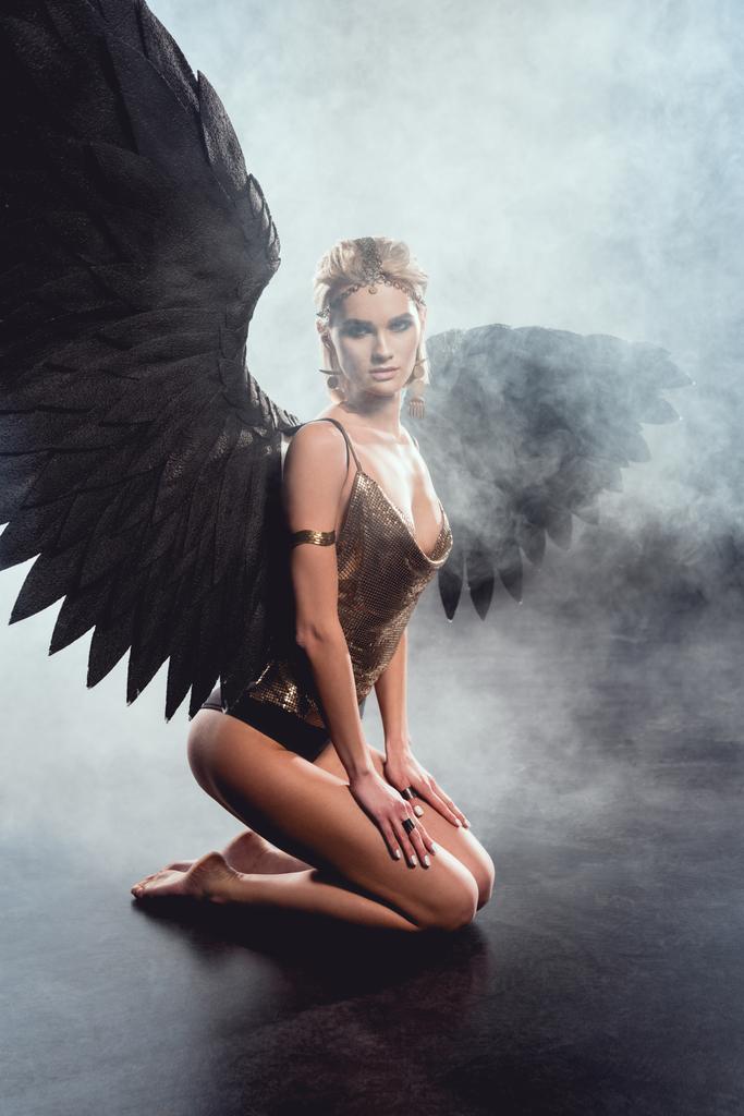 szép szexi nő ül, látszó-on fényképezőgép, és pózol a sötét háttér előtt fekete angyal szárnyakkal - Fotó, kép