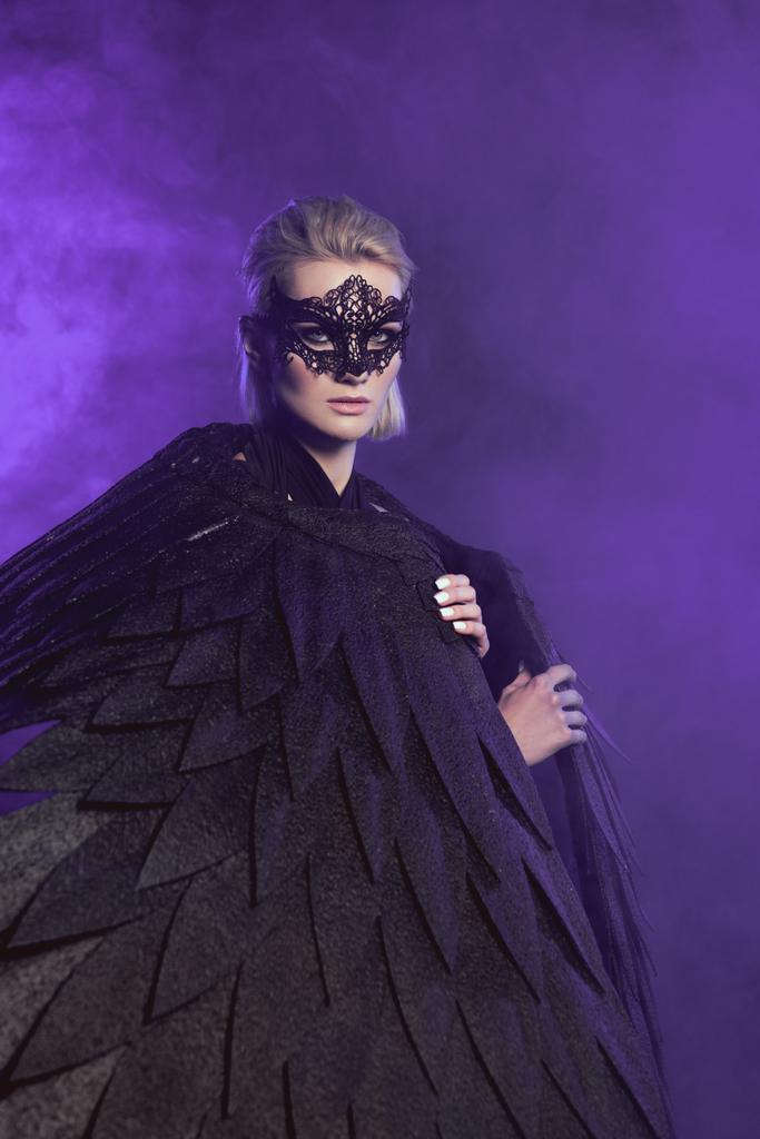 krásná žena v krajkové masky a černá andělská křídla při pohledu na fotoaparát a Pózování na fialové pozadí - Fotografie, Obrázek