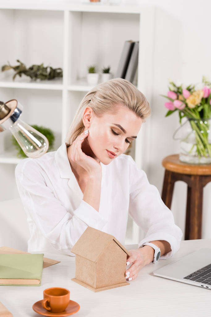 attractive woman looking at small cardboard house while sitting near cup with drink, environmental saving concept  - Photo, Image