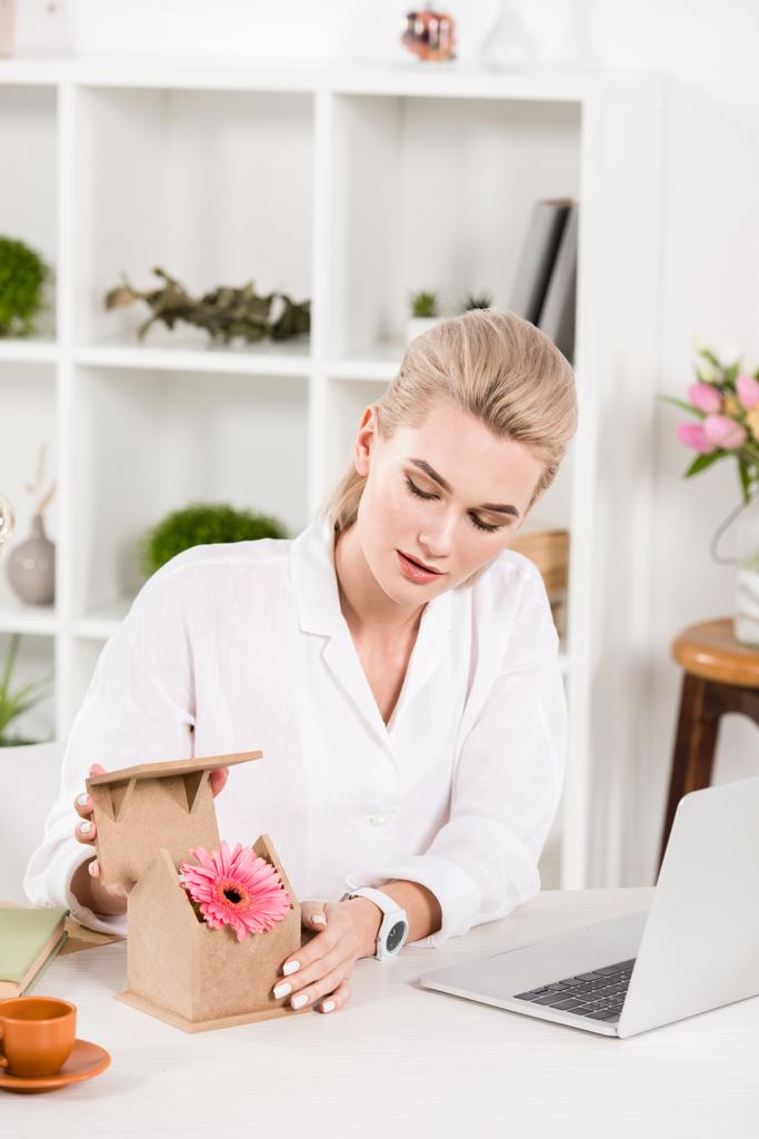 attractive woman looking at pink flower in small cardboard house while sitting near laptop in office, environmental saving concept  - Photo, Image
