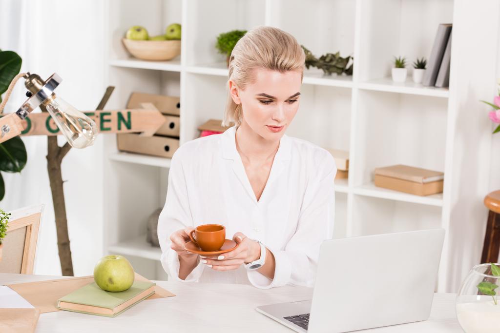 atractiva mujer mirando a la computadora portátil mientras sostiene la taza en la oficina
  - Foto, Imagen