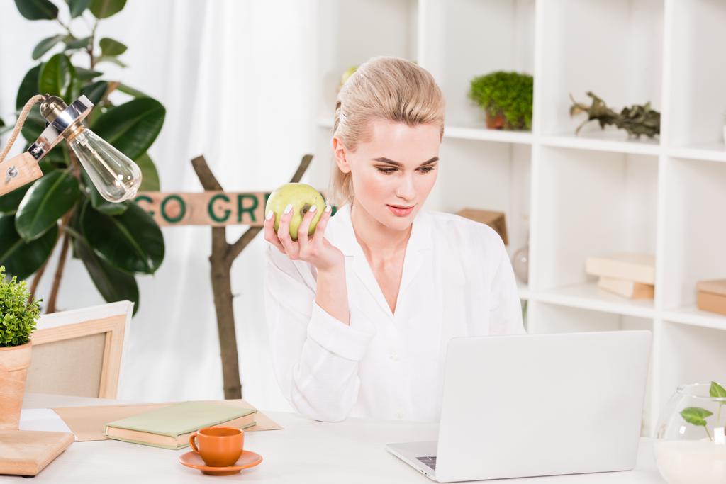 houkutteleva nainen katselee kannettavaa tietokonetta ja pitää vihreää omenaa toimistossa
  - Valokuva, kuva
