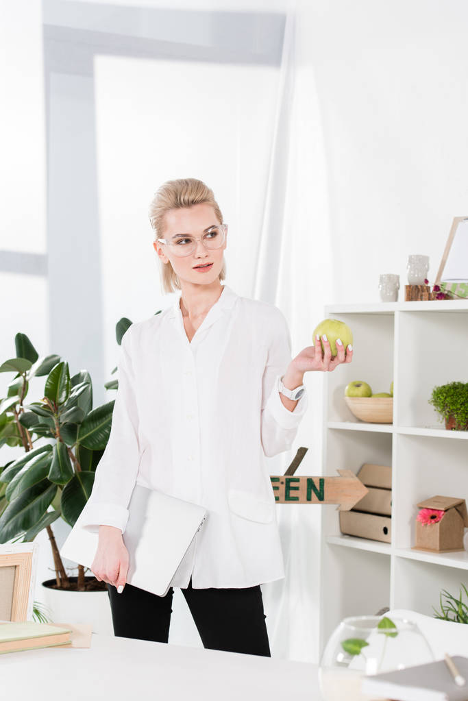 attractive woman standing with laptop and holding green apple in office  - Photo, Image