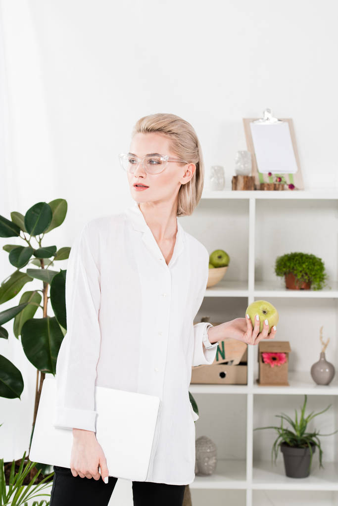 bella donna in occhiali in piedi con computer portatile e mela verde in ufficio
  - Foto, immagini