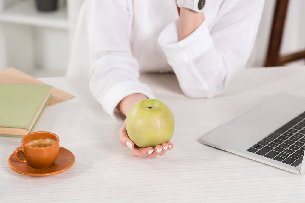 cropped view of woman holding green apple near laptop and cup in office  - Photo, Image