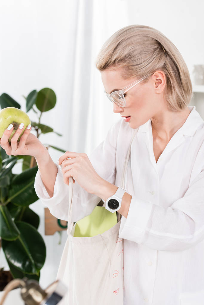 hermosa mujer de negocios en gafas con manzana verde y mirando dentro de la bolsa ecológica, concepto de ahorro ambiental
  - Foto, imagen