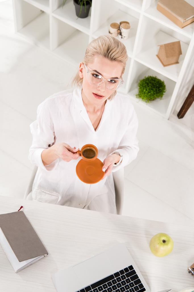 overhead view of woman in glasses holding cup of coffee near desk in office - Photo, Image