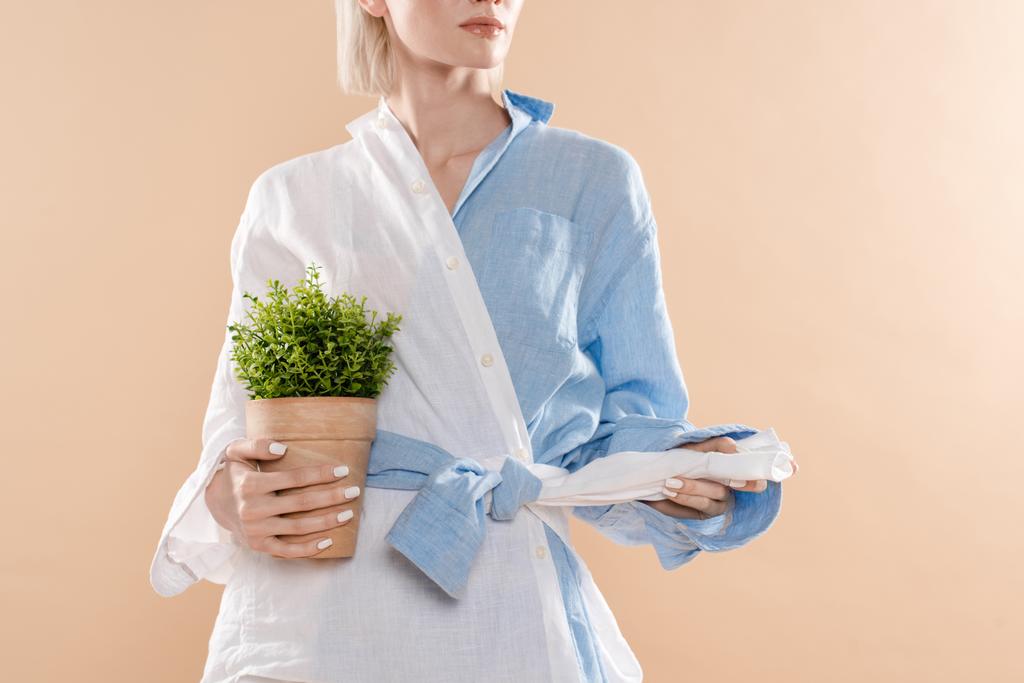 περικοπεί άποψη της γυναίκας κρατώντας το δοχείο με το φυτό και στέκεται σε οικολογικά ρούχα απομονώνονται σε μπεζ, περιβαλλοντική εξοικονόμηση έννοια  - Φωτογραφία, εικόνα