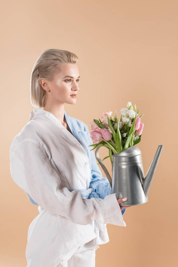 mooie vrouw houdt van gieter met bloemen en permanent in eco kleding geïsoleerd op beige, milieu opslaan concept  - Foto, afbeelding