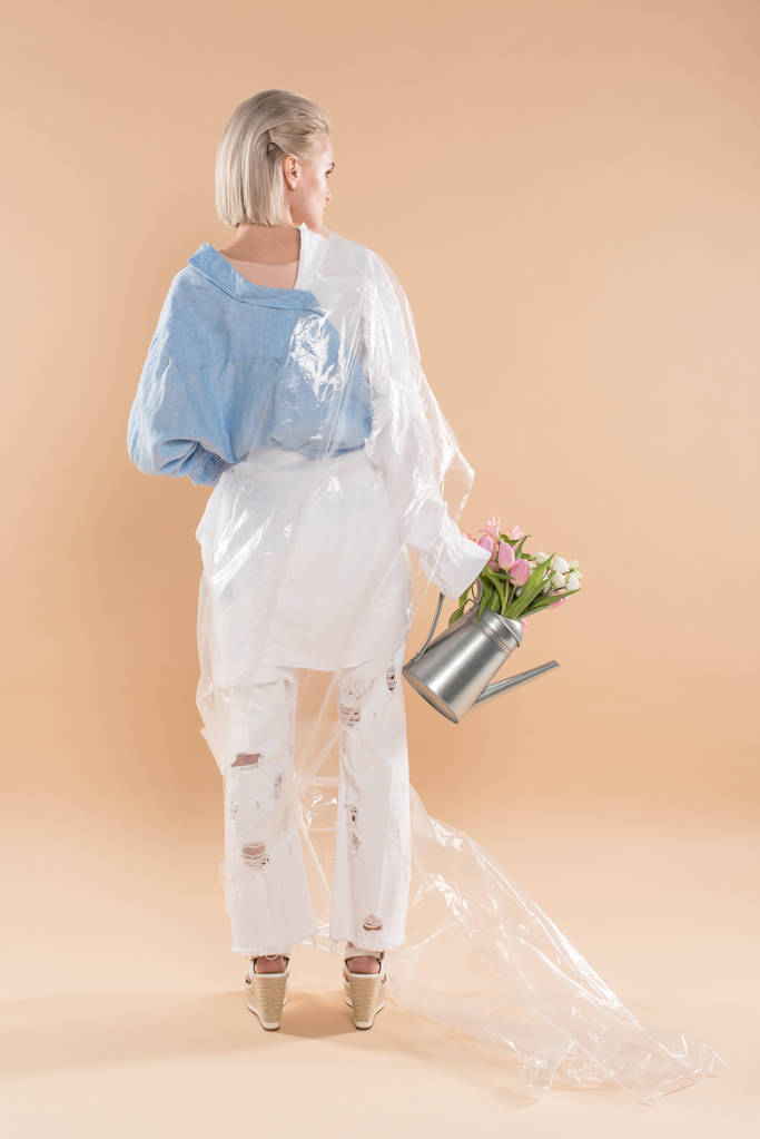 κορίτσι κρατώντας ποτιστήρι με λουλούδια και στέκεται σε οικολογικά ρούχα τυλιγμένο σε πολυαιθυλένιο που απομονώνονται σε μπεζ, περιβαλλοντική σωτήρια ιδέα  - Φωτογραφία, εικόνα