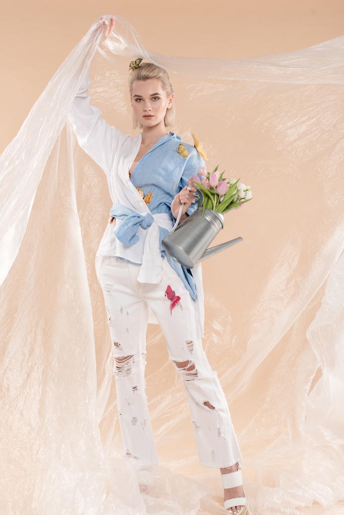 donna in possesso di polietilene e annaffiatoio con fiori mentre in piedi in eco abbigliamento isolato su beige, concetto di risparmio ambientale
  - Foto, immagini