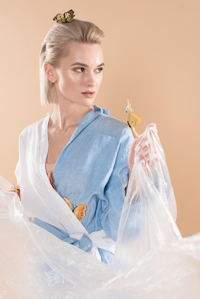 attractive girl holding polyethylene while standing with butterflies on eco clothing isolated on beige, environmental saving concept  - Photo, Image