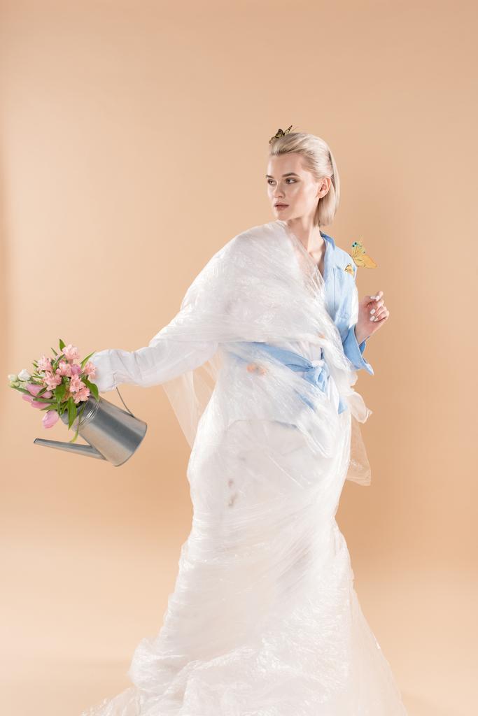 krásná dívka v polyethylenu drží konev s květinami a stojí v oděv eco samostatný na béžové, ekologické ukládání konceptu  - Fotografie, Obrázek