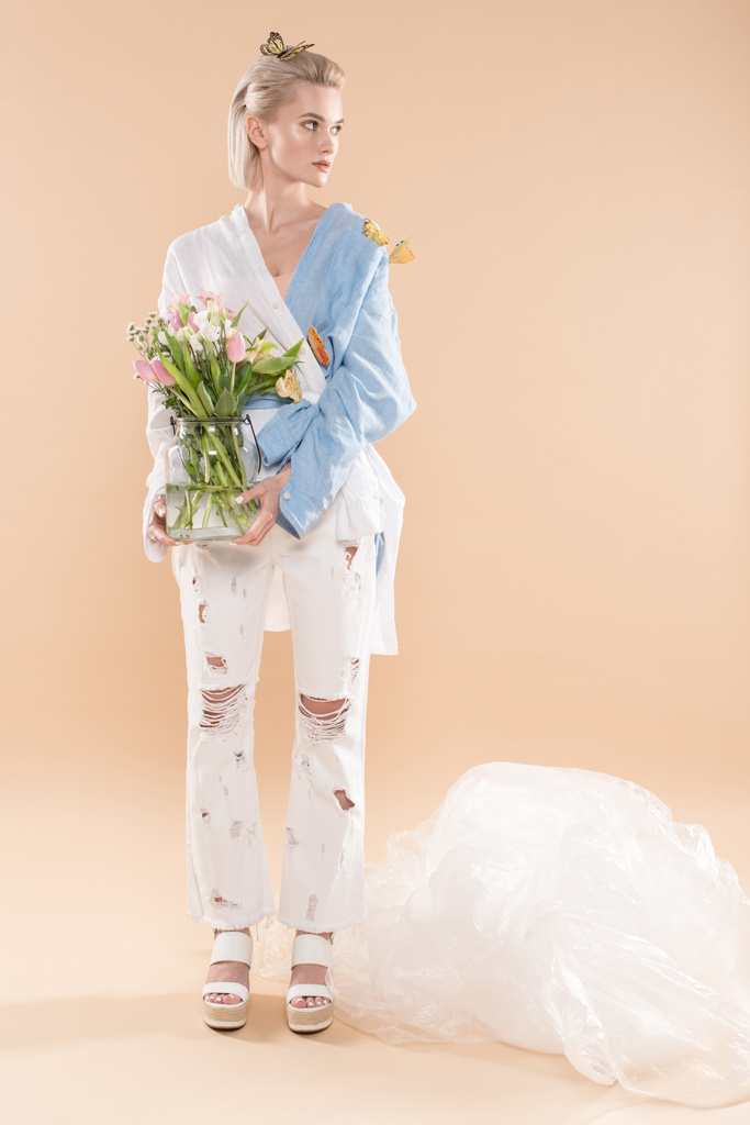 beautiful yound woman standing with butterflies on eco clothing and holding glass vase with flowers on beige background, environmental saving concept - Photo, Image
