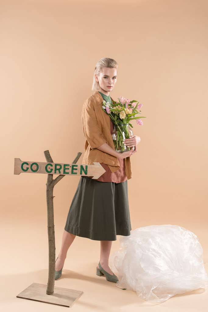 attractive girl in eco clothing holding vase with flowers near sign with go green lettering on beige background, environmental saving concept - Photo, Image
