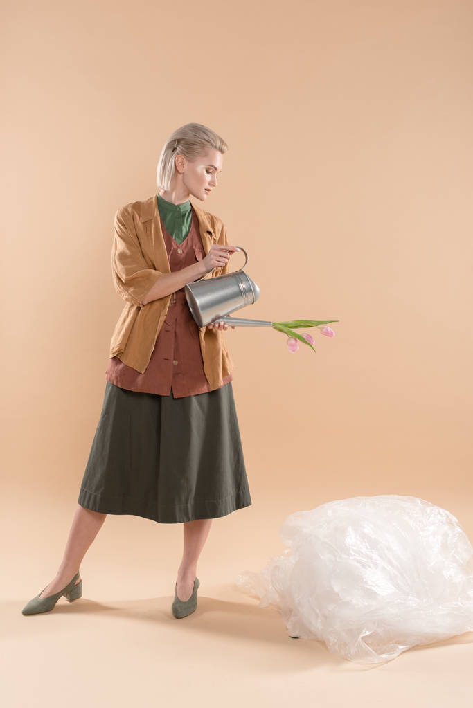 ξανθιά κοπέλα σε οικολογικά ρούχα κρατώντας ποτιστήρι με λουλούδια κοντά πολυαιθυλενίου σε μπεζ φόντο, περιβαλλοντική εξοικονόμηση έννοια - Φωτογραφία, εικόνα