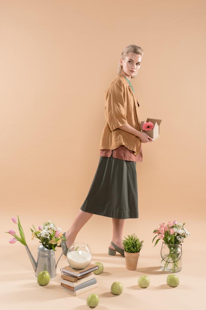atrakcyjna kobieta trzyma kartonowy Domek z kwiat w pobliżu rośliny i kwiaty na beżowym tle, środowiska zapisywanie koncepcja - Zdjęcie, obraz
