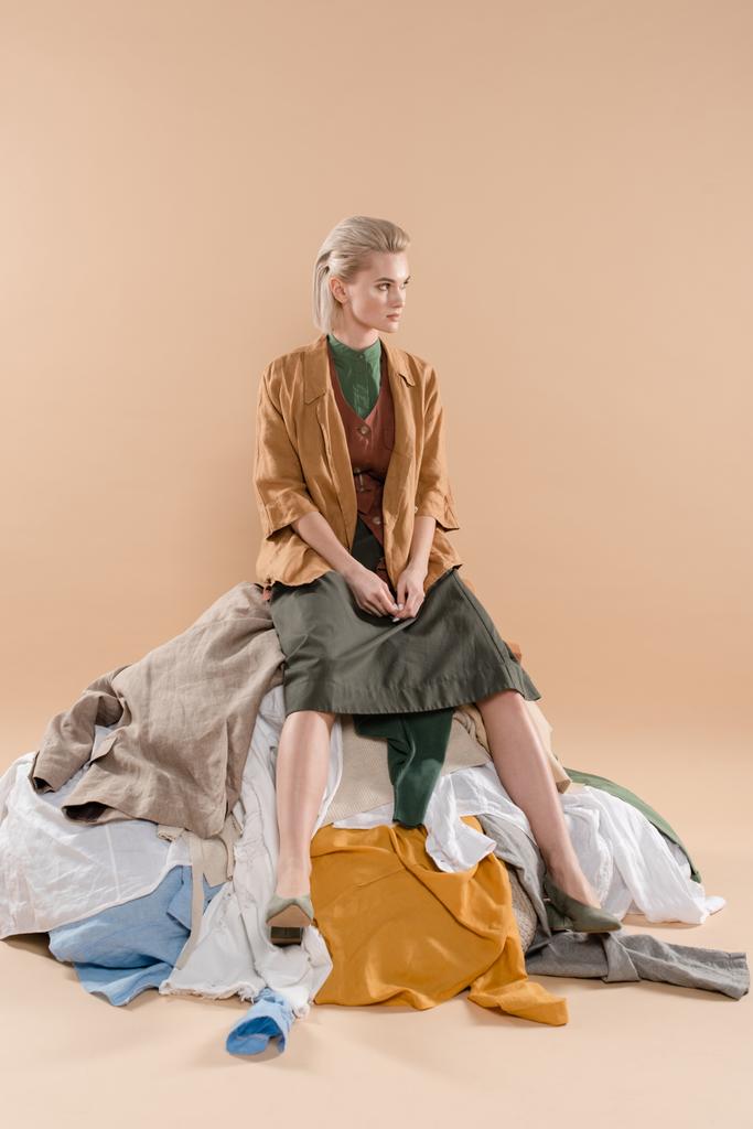 mulher loira sentada na pilha de roupas em fundo bege, conceito de economia ambiental
 - Foto, Imagem