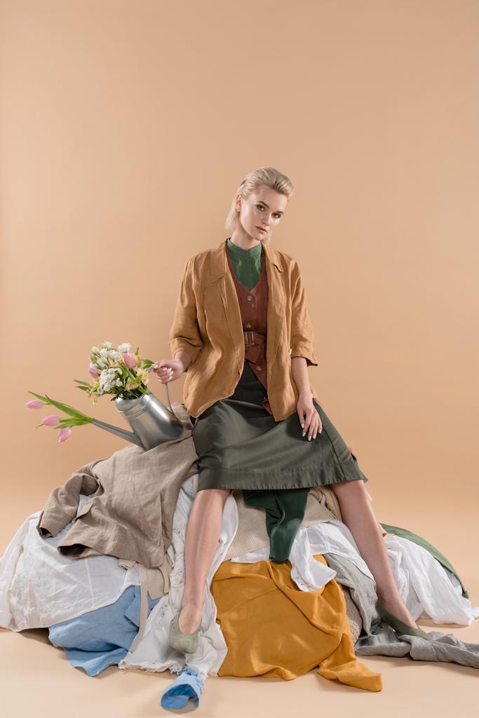 chica rubia sentada en la pila de ropa y sosteniendo regadera con flores sobre fondo beige, concepto de ahorro ambiental
 - Foto, Imagen
