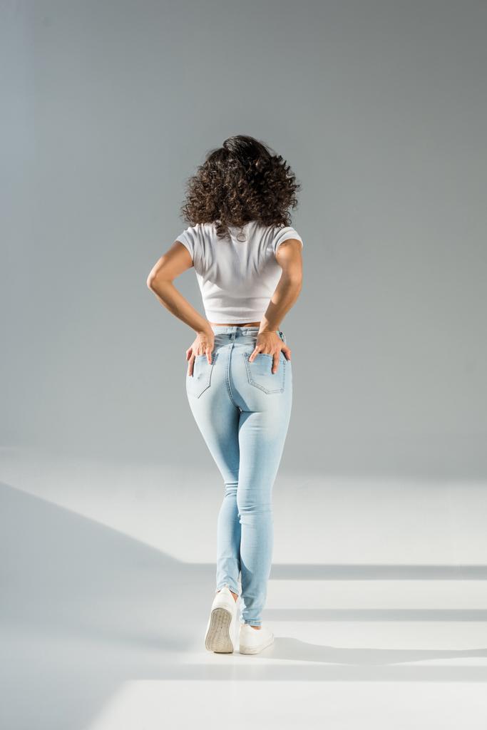 Widok z kobieta stojąc z rękami w kieszeniach w obcisłe niebieskie dżinsy na szarym tle tyłu - Zdjęcie, obraz